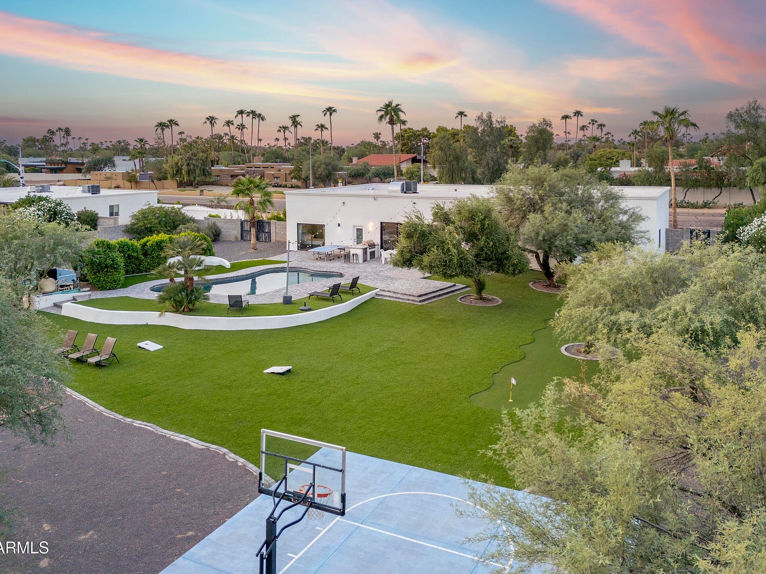 Aerial view of the property at twilight showing the full backyard: pool, pickleball court, basketball half-court, sand volleyball area, putting green, and the long single-story ranch behind, framed by palm trees