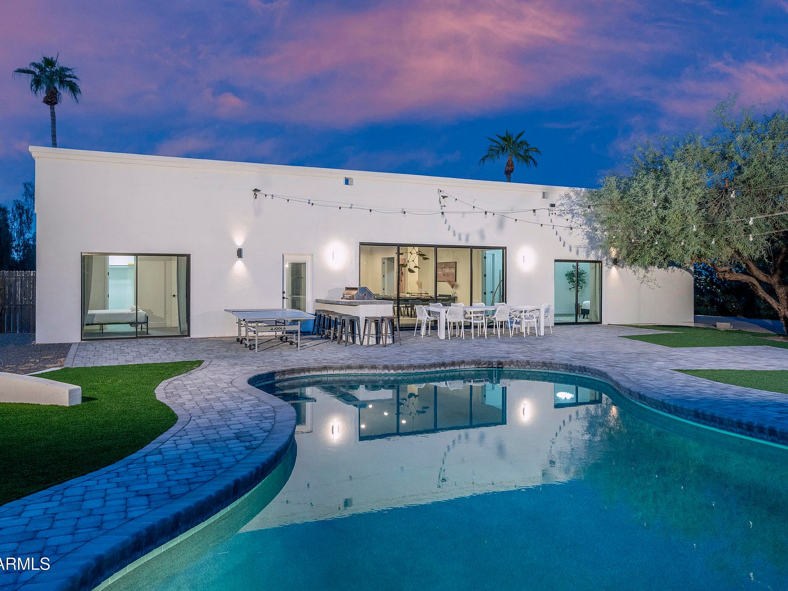 Twilight rear elevation of the house seen across the curved pool, string lights, a ping-pong table on the paver patio, dining set under the eave, glass sliders open to the lit interior