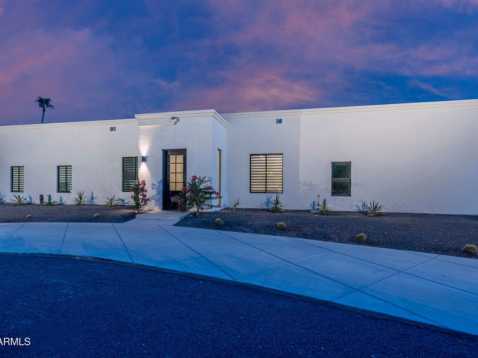 Front elevation at dusk: low single-story modern stucco facade with a centered black-framed front door, lit sconces, agave and cactus landscape, deep pink and purple Sonoran sky overhead
