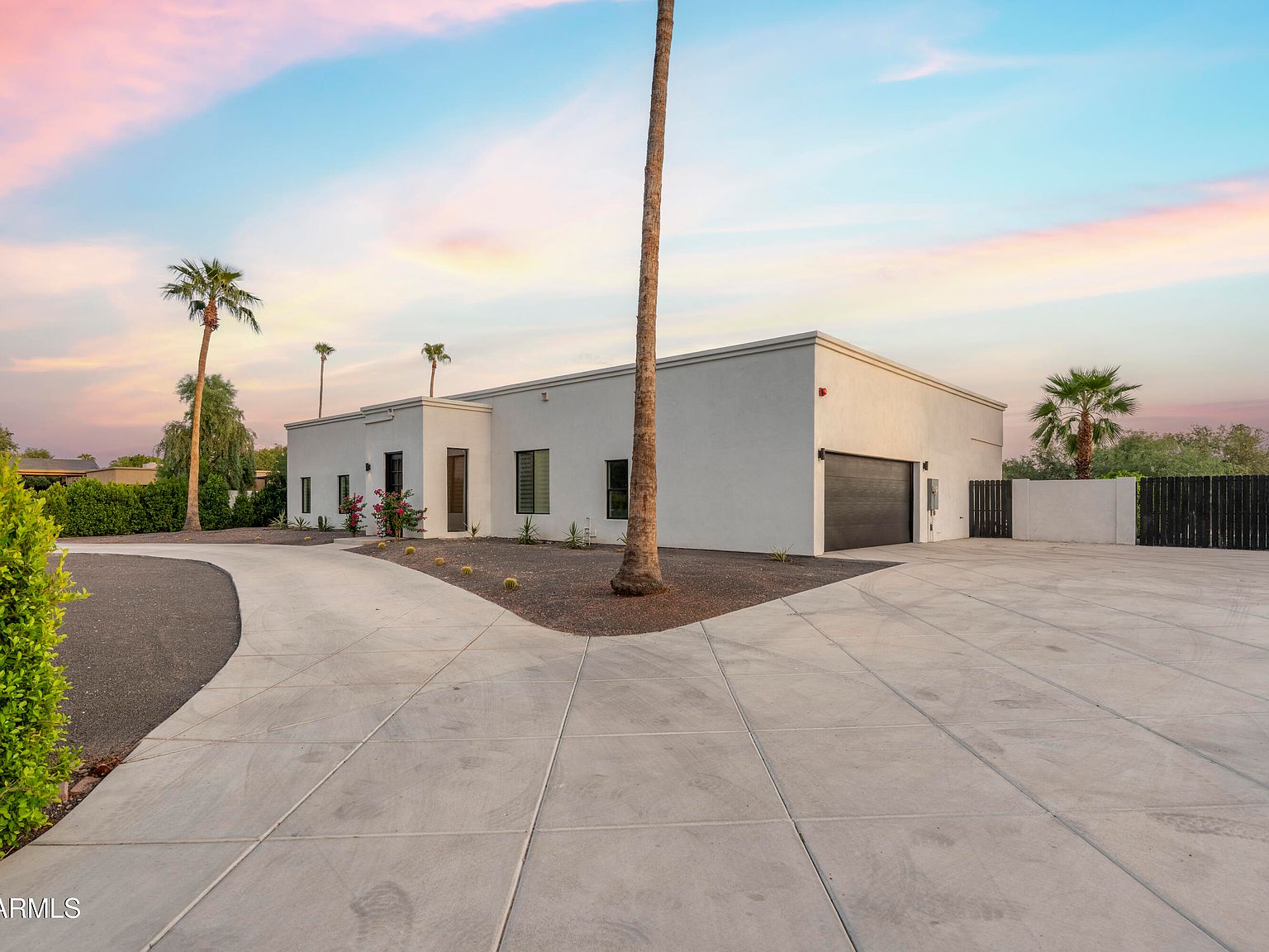 Side approach at dusk showing a mature palm anchoring a wide paver driveway, the dark garage door and RV gate at right, the long flat-roof ranch stretching back across the lot