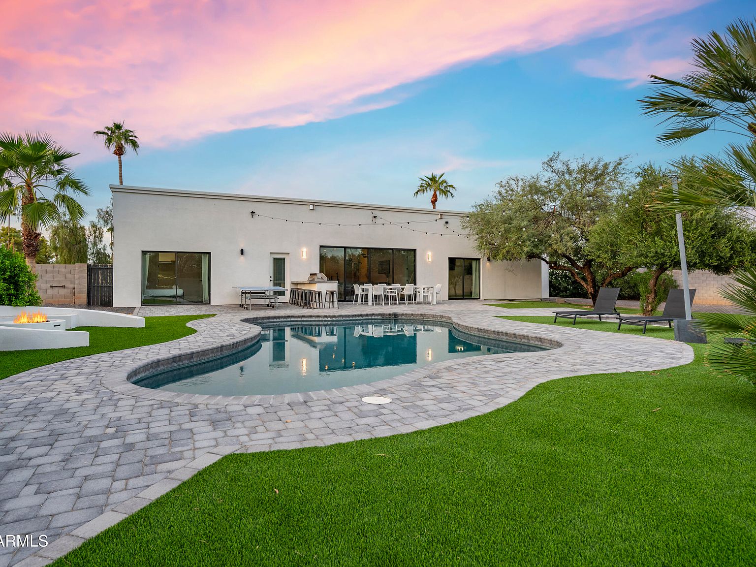 Twilight view of 8417 E Cactus Rd in Scottsdale: a long modern ranch with walls of glass, a curved pool in the foreground, fire pit and lounge chairs on a paver patio, palm trees against a pink-peach Arizona evening sky